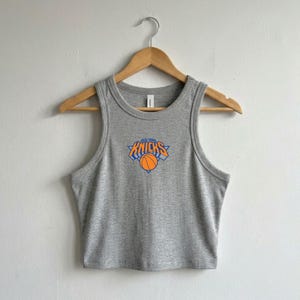 May include: A heather grey sleeveless crop top featuring a ribbed crew neckline and armholes. The centre of the chest displays the New York Knicks logo, which includes the team name in orange text and a basketball graphic inside a blue triangle.