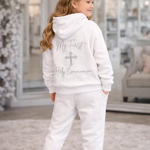 Puede incluir: Conjunto blanco de sudadera con capucha y pantalón de chándal a juego. La parte trasera de la sudadera con capucha presenta el texto "My First Holy Communion" con un diseño de cruz. El conjunto se combina con zapatillas de purpurina plateadas y una diadema floral.