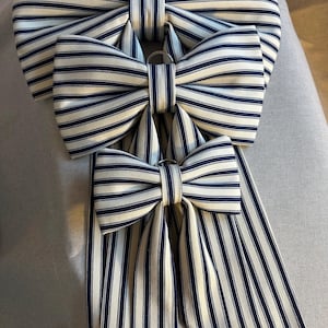 May include: Three striped bows in varying sizes, arranged vertically. The bows feature a cream-colored fabric with navy blue stripes. Each bow has a metal ring at the center. The bows are likely decorative accessories.