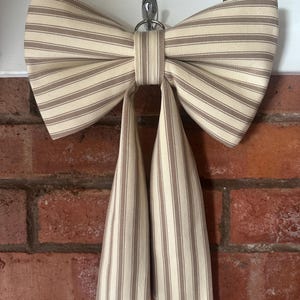 May include: A decorative fabric bow featuring a cream and tan vertical striped pattern. The bow has two wide, pleated loops and two long, straight hanging tails. It is constructed from a woven textile material and is suspended from a metal hook.