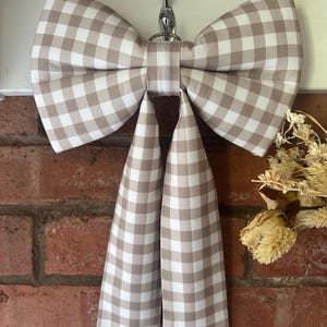 Op de afbeelding: Een decoratieve strik van stof met een beige en wit gingham ruitpatroon. De strik heeft twee grote lussen en twee lange, rechte hangende uiteinden, met een centraal knoopdetail en een metalen ring aan de bovenkant om de strik op te hangen.