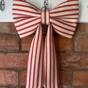 May include: A decorative bow made of fabric featuring a pattern of vertical red and cream-coloured stripes. The bow has two wide loops and two long, hanging tails that extend downward. A small metal ring is attached to the centre knot of the bow.