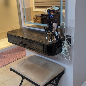May include: A modern vanity set with a dark wood floating shelf, a lighted mirror, and a cushioned stool. The shelf has a drawer and holds hair styling tools. The stool has a black metal frame and a beige cushion.
