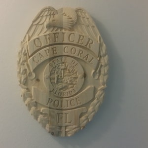 May include: A wooden plaque shaped like a police badge with the text "OFFICER CAPE CORAL STATE OF FLORIDA POLICE FL" carved into it.