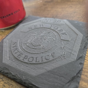 May include: A dark gray slate coaster with the New York State Police emblem etched into the surface. The emblem includes the words "NYSP", "New York", and "State Police". A red can with the words "Calorie Cola" is partially visible.