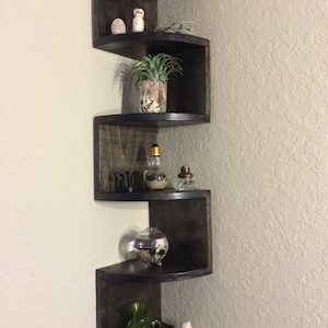 May include: A set of five black corner shelves with rounded edges. The shelves are stacked in a staggered pattern and feature various decorative items, including plants, a glass globe, and small figurines.