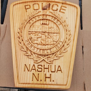 May include: A wooden plaque with the words "Police" and "Nashua N.H." carved into it. The plaque also features a circular emblem with the words "Nashua 1853 Dunstable 1673" and a depiction of a train crossing a bridge.