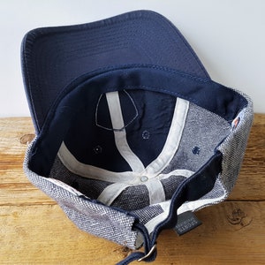 May include: A navy blue baseball cap with a curved brim. The interior features white support bands and a dark blue sweatband. The cap has a gray and white speckled fabric on the sides and a small tag that reads "SPORTS SPECIALTIES."