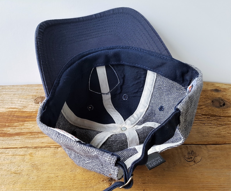 May include: A navy blue baseball cap with a curved brim. The interior features white support bands and a dark blue sweatband. The cap has a gray and white speckled fabric on the sides and a small tag that reads "SPORTS SPECIALTIES."
