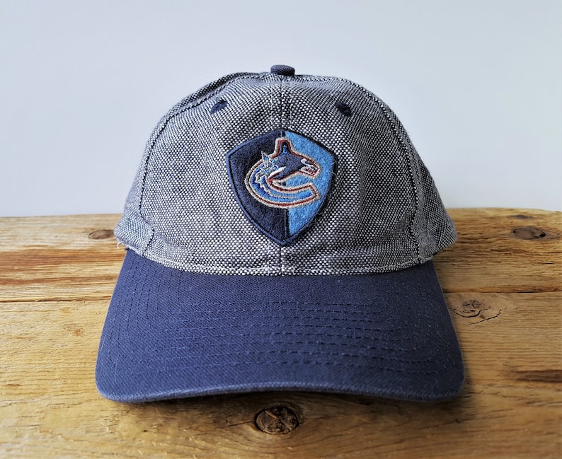 May include: A blue and gray baseball cap with the Vancouver Canucks logo on the front. The cap has a curved brim and a fabric texture. The logo is a shield shape with a stylized orca.