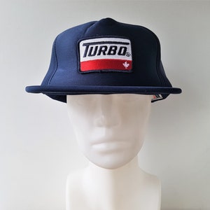 May include: Navy blue flat-brim hat with a rectangular patch featuring the word "TURBO" in white, a red stripe, and a small maple leaf. The hat is displayed on a white mannequin head.