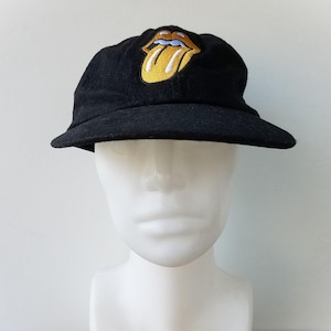 May include: Black baseball cap featuring a yellow and white embroidered tongue logo. The cap has a curved peak and is displayed on a white mannequin head. A classic hat.