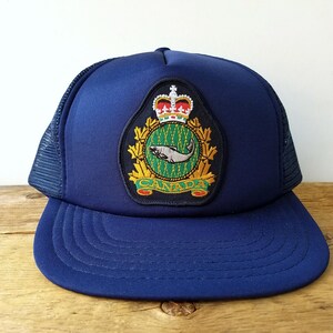May include: A navy blue trucker hat with a curved peak and mesh back. The hat features a patch with a crown, a fish, and the word "CANADA" in gold lettering. The patch is surrounded by a green and gold design.