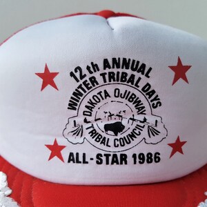 May include: Red and white trucker hat with the text "12th Annual Winter Tribal Days Dakota Ojibway Tribal Council All-Star 1986". The hat features red stars and a mesh back.