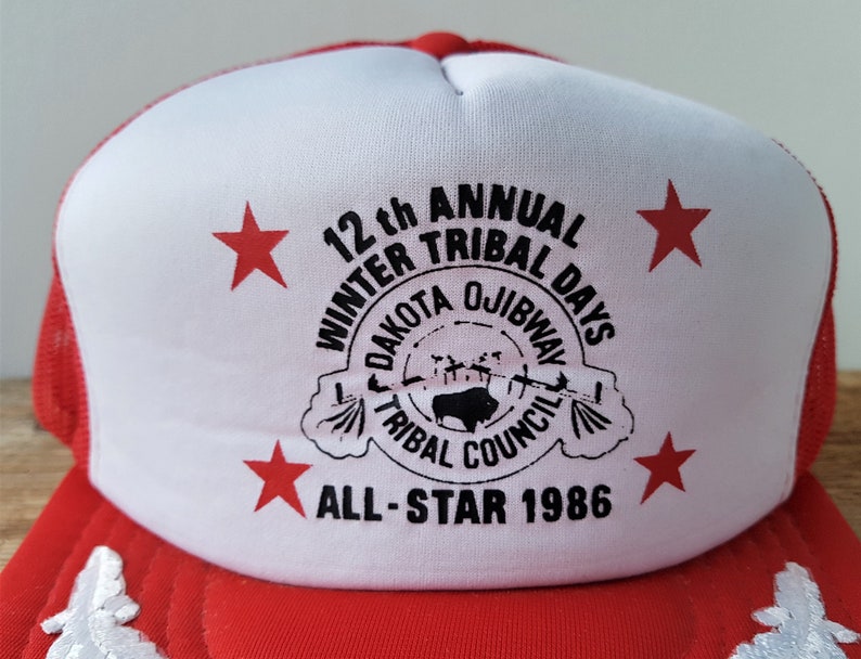 May include: Red and white trucker hat with the text "12th Annual Winter Tribal Days Dakota Ojibway Tribal Council All-Star 1986". The hat features red stars and a mesh back.