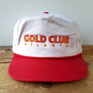 May include: A white and red baseball cap featuring the embroidered text "GOLD CLUB ATLANTA" in orange. The cap has a red peak and a red rope detail along the base of the white crown.