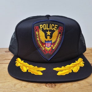 May include: Black trucker cap with a mesh back and a flat peak. The front displays a police badge with the text "POLICE" and "CITY OF CORINTH MISSISSIPPI". Gold leaf embroidery decorates the peak.