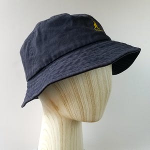 K&ouml;nnte beinhalten: Ein marineblauer Fischerhut mit leicht abgenutztem Aussehen. Der Hut hat ein kleines, gesticktes gelbes K&auml;nguru-Logo auf der Vorderseite. Der Hut wird auf einem h&ouml;lzernen Schaufensterpuppenkopf pr&auml;sentiert.