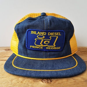 Może przedstawiać: Niebieska i żółta czapka trucker z naszywką z napisem "Inland Diesel id Prince George".