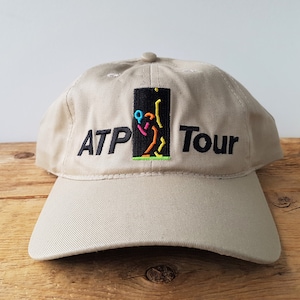 Op de afbeelding: Beige baseballpet met de geborduurde tekst "ATP Tour". De pet heeft een kleurrijke afbeelding van een tennisser. De hoed heeft een gebogen klep en een knoop aan de bovenkant.