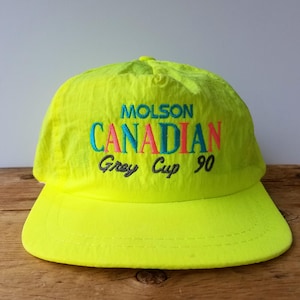 Puede incluir: Gorra de béisbol amarilla neón con visera curva. La gorra presenta texto bordado en turquesa, rosa y negro que dice "Molson Canadian Grey Cup 90". La gorra está hecha de un material ligero, posiblemente nailon.