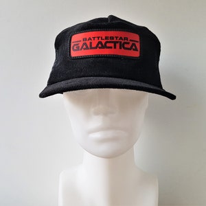 Puede incluir: Una gorra de béisbol de pana negra con un parche rojo que dice "Battlestar Galactica" en letras blancas.
