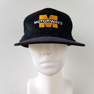 May include: Black corduroy hat with a flat brim. The hat features the word "MOTORWAYS" embroidered in white over a gold "M" logo. The hat is displayed on a white mannequin head.