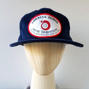 May include: A navy blue corduroy cap with a curved brim. The cap features a white and red oval patch with the text "CORNELL PUMPS" and "WAYNE GILBERTSON SALES & SERVICE LTD. BOW ISLAND". The patch includes a graphic of a red and white wheel.