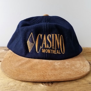 Op de afbeelding: Een marineblauwe baseballpet met een beige suède klep. De voorzijde is voorzien van goudkleurig borduurwerk met de tekst CASINO de MONTRÉAL en een ruitvormig logo. De pet heeft een gestructureerde kroon en een knoop aan de bovenzijde.