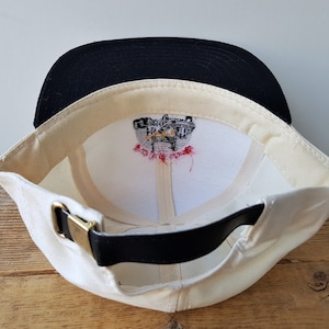 Op de afbeelding: Cr&egrave;mekleurige baseballpet met een zwarte klep. De binnenkant heeft een witte voering met een geborduurd ontwerp en een zwarte leren riem met een goudkleurige gesp. De pet is schuin geplaatst om de binnenkant te laten zien.