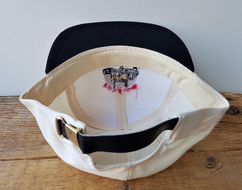 Op de afbeelding: Cr&egrave;mekleurige baseballpet met een zwarte klep. De binnenkant heeft een witte voering met een geborduurd ontwerp en een zwarte leren riem met een goudkleurige gesp. De pet is schuin geplaatst om de binnenkant te laten zien.