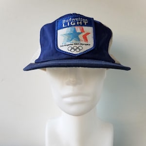Könnte beinhalten: Eine blaue und weiße Trucker-Kappe mit einem Budweiser Light-Logo und dem Text "Los Angeles 1984 Olympics". Das Logo zeigt einen rot-weiß-blauen Stern mit einem stilisierten amerikanischen Flaggen-Design.