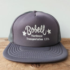 May include: Grey trucker hat with a white embroidered logo that says "Bobell Northwest Transportation LTD." and two white stars.