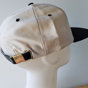 Op de afbeelding: Een beige baseballpet met een zwarte klep en zwarte stiksels. De pet heeft een zwarte leren band met een goudkleurige metalen gesp voor verstelling. De pet wordt getoond op een witte mannequinhoofd.