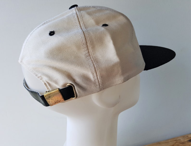 Op de afbeelding: Een beige baseballpet met een zwarte klep en zwarte stiksels. De pet heeft een zwarte leren band met een goudkleurige metalen gesp voor verstelling. De pet wordt getoond op een witte mannequinhoofd.