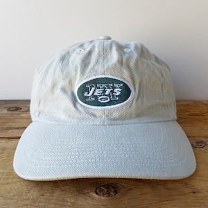 May include: A light beige baseball cap featuring an embroidered oval patch on the front. The patch has a dark green background with the white text JETS and a small football graphic in the centre. The cap has a curved brim and stitched eyelets.