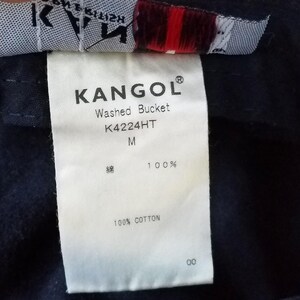 K&ouml;nnte beinhalten: Nahaufnahme eines wei&szlig;en Etiketts mit dem Markennamen "KANGOL" in Schwarz aufgedruckt. Das Etikett enth&auml;lt auch den Text "Washed Bucket", "K4224HT" und "M". Das Etikett gibt an, dass der Hut aus 100 % Baumwolle besteht.