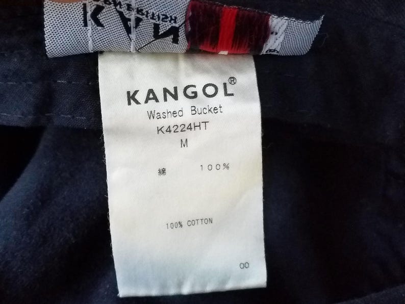 K&ouml;nnte beinhalten: Nahaufnahme eines wei&szlig;en Etiketts mit dem Markennamen "KANGOL" in Schwarz aufgedruckt. Das Etikett enth&auml;lt auch den Text "Washed Bucket", "K4224HT" und "M". Das Etikett gibt an, dass der Hut aus 100 % Baumwolle besteht.