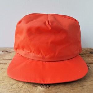May include: Bright orange cap with a rounded crown and a curved brim. The cap is made of a smooth, possibly nylon or polyester fabric. The brim is a solid, matching orange color.