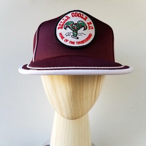 Könnte beinhalten: Burgunderfarbene Baseballkappe mit weißem Rand und Zierleiste. Die Kappe hat einen runden Aufnäher mit dem Text "BELLA COOLA B.C. HOME OF THE THUNDERBIRD" und einer Grafik eines Thunderbirds. Die Kappe wird auf einem hölzernen Schaufensterpuppenkopf präsentiert.