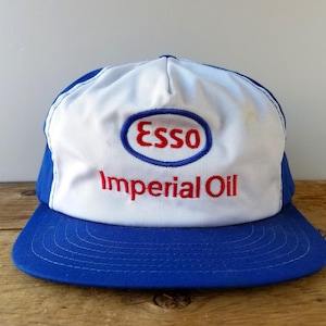 May include: A vintage Esso Imperial Oil baseball cap. The cap features a white front panel with the Esso logo in a blue oval and red text. The brim and back are blue.