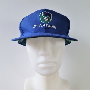 Op de afbeelding: Een blauwe baseballpet met een groen en wit honkbalhandschoenlogo en de tekst "ST-ANTOINE" geborduurd op de voorkant.