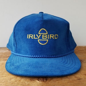 May include: A blue corduroy baseball cap with a flat peak and a rope detail. The cap features the embroidered text "IRLY BIRD" in gold. The hat has a classic trucker hat design.