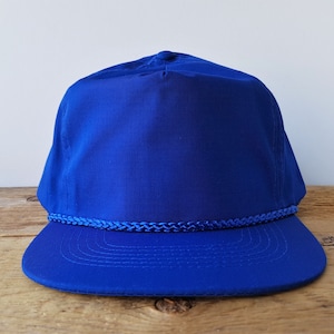 May include: A solid blue baseball cap with a rope trim around the crown. The cap has a curved brim.