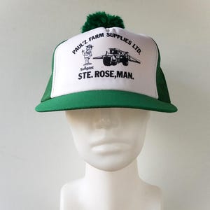 以下が含まれることがあります： 「PAUL'Z FARM SUPPLIES LTD.」と「STE. ROSE, MAN.」の文字が入った、緑と白のトラッカーハット。つばは緑色、フロントパネルはグラフィック入りの白色、バックは緑色のメッシュ。上部には緑色のポンポンが付いています。