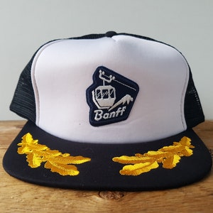 May include: White and black trucker hat with a navy blue embroidered patch featuring a ski lift and the word "Banff". The brim of the hat is black with gold embroidered leaves.