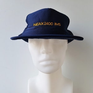Op de afbeelding: Marineblauwe vissershoed met de tekst "NEAX 2400 IMS" in goud geborduurd. De hoed heeft een mesh gedeelte en een rand. De hoed wordt getoond op een witte mannequin hoofd.