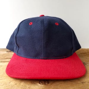 Peut inclure: Casquette de baseball bleu marine avec une visière rouge. Le chapeau a un design classique avec une couronne structurée et une visière incurvée. La casquette a des coutures rouges et un bouton rouge sur le dessus.