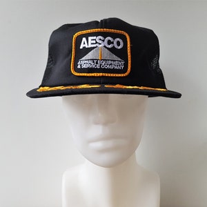 May include: Black mesh trucker hat with a yellow and white embroidered patch that says "AESCO Asphalt Equipment & Service Company".