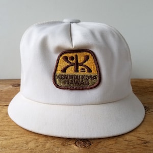 Pode incluir: Boné de beisebol branco com aba curva. O boné apresenta um patch retangular com as palavras "KEAUHOU KONA HAWAII" em letras marrons. O patch tem um fundo amarelo e verde com uma borda marrom.